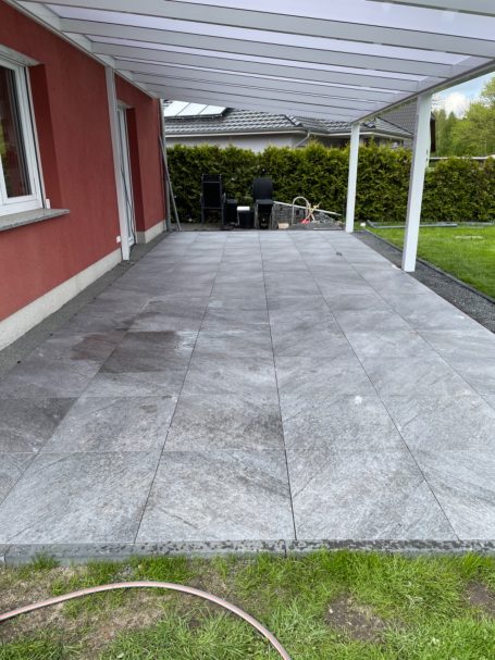 Terrasse mit grauen Fliesen, überdacht, an einem roten Haus. Rasen im Hintergrund.